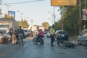 Eine Gruppe von Menschen steht um ein verunglücktes Motorrad auf der Straße mit mehreren Fahrzeugen, darunter ein Lastwagen, und einer Hintergrund von Bäumen, Pfählen, Lampen und Schildern unter dem Himmel.