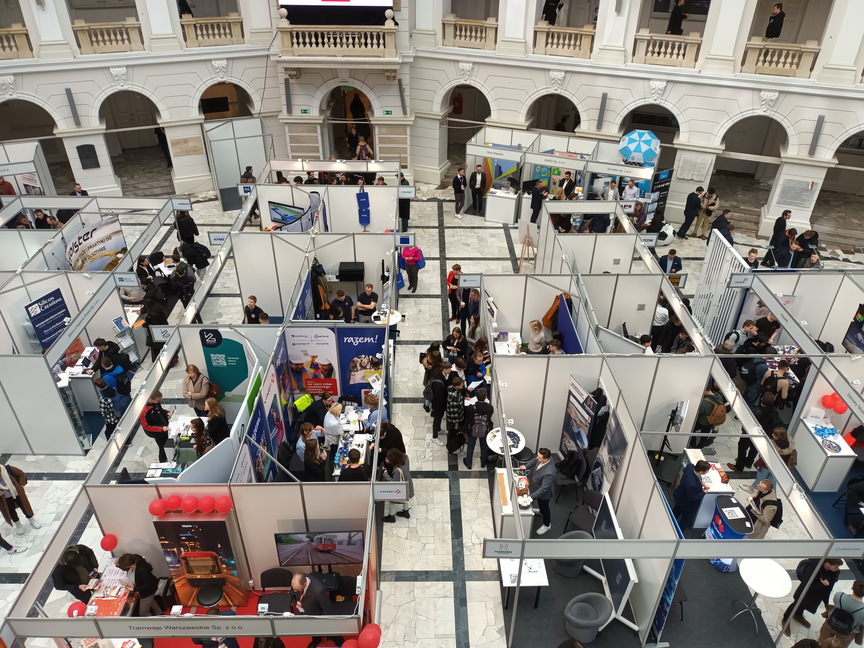 Eine große Gruppe von Menschen auf einer internationalen Fachmesse in einem Raum mit Ständen, Bannern, Tischen, Stühlen, Luftballons und einem Gebäude mit Bögen, Säulen, Geländern und einem Bildschirm im Hintergrund.