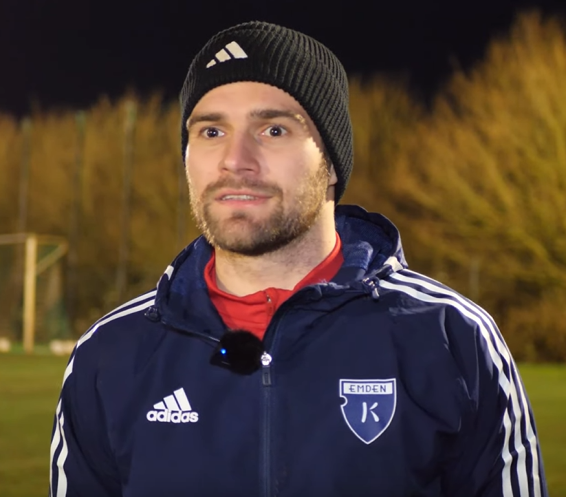 Ein Mann in einer blauen Jacke und einem Hut steht auf einem Fußballfeld, umgeben von Bäumen und einem Tor im Hintergrund während eines Trainings.