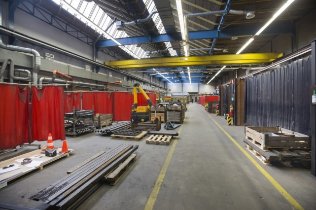 Großes Lagerhausinneres mit Holzpaletten, Metallobjekten, Verkehrskegeln und Maschinenfahrzeug, mit roten Vorhängen, Rohren, Deckenlampen und einer verhängten Wand.