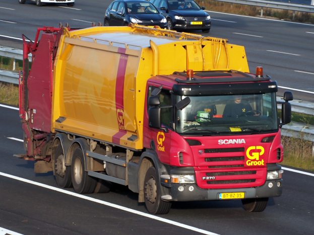 Ein gelb-roter Müllwagen fährt auf einer Autobahn, umgeben von anderen Fahrzeugen, mit Gras und einer Leitplanke auf beiden Seiten der Straße.