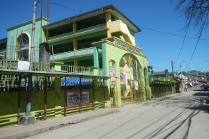Ein grünes Gebäude an einer Straßenecke mit Strommasten, Kabeln, Bäumen, Fahrzeugen und Passanten unter einem sichtbaren Himmel.