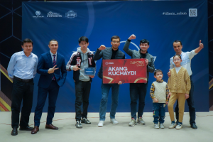 Eine Gruppe von Menschen auf einer Bühne, zwei halten ein Schild mit der Aufschrift "Akang Kuchaydi - Zakovat Quiz", mit einem Banner, Scheinwerfern und anderen Gegenständen im Hintergrund.