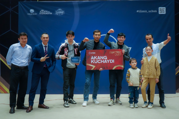 Eine Gruppe von Menschen auf einer Bühne, zwei halten ein Schild mit der Aufschrift "Akang Kuchaydi - Zakovat Quiz", mit einem Banner, Scheinwerfern und anderen Gegenständen im Hintergrund.