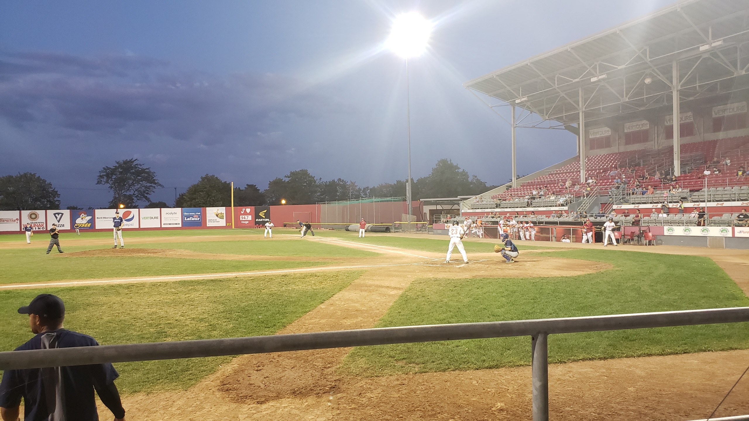 Baseballspiel im Gange mit Zuschauern auf den Tribünen, Geländer im Vordergrund, Trees und Stadion-Infrastruktur sichtbar unter einem klaren blauen Himmel.