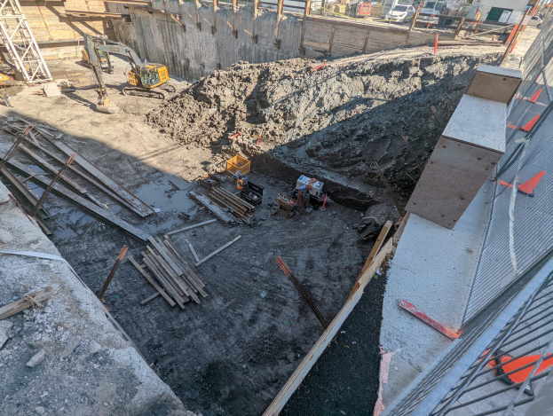 Baustelle mit einem großen Schutthaufen, Kränen, Holzstangen, einem Maschendrahtzaun auf der rechten Seite und Gebäuden, Fahrzeugen, Polen und Bäumen im Hintergrund unter dem Himmel.