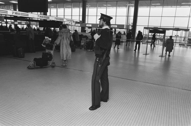 Ein Polizeibeamter in Uniform steht in einem Flughafen-Terminal, hält eine Waffe und ist von Menschen und Gepäck umgeben, mit Gepäckwagen, Deckenleuchten, Schildern, Pfosten und Glaswänden im Hintergrund.
