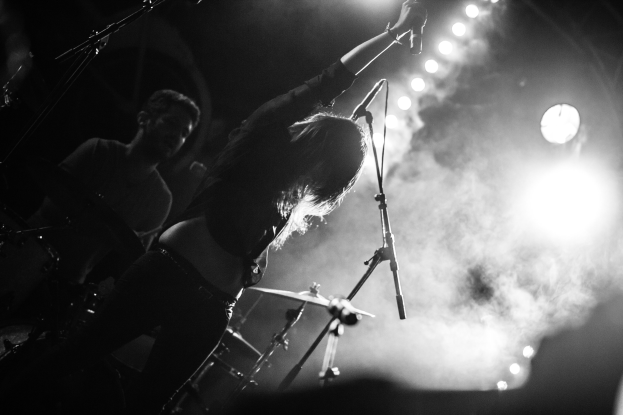 Schwarz-weiß-Foto einer Frau, die auf der Bühne in ein Mikrofon singt, mit einem Schlagzeuger im Hintergrund und Bühnenbeleuchtung.