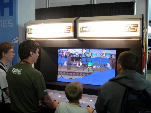 Ein Junge spielt ein Videospiel, während drei Männer stehen und zusehen; ein Vorhang und ein blaues Schild an einer Säule sind im Hintergrund.