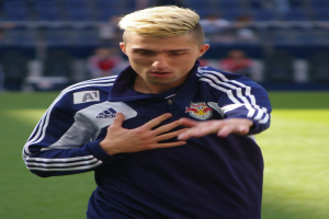 Ein junger Mann in einem blauen T-Shirt steht auf einem Fußballfeld und streckt die Arme aus, mit einem entschlossenen Gesichtsausdruck.