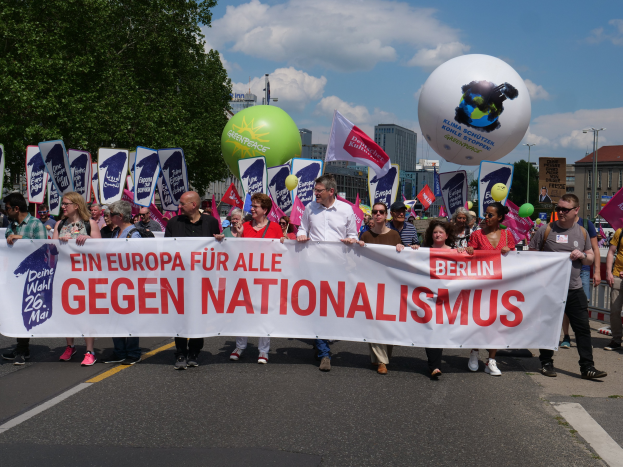 Eine Gruppe von Menschen marschiert auf einer Straße in Berlin, hält ein Transparent, Fahnen und Luftballons, mit Bäumen und Gebäuden auf der linken Seite und einem bewölkten Himmel im Hintergrund, nimmt an einer Demonstration gegen Nationalismus teil.