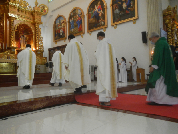 Eine Gruppe von Priestern geht einen roten Teppich in einer Kirche entlang, mit Menschen, die auf dem Boden vor und rechts von ihnen stehen, und Foto Rahmen an der Hintergrundwand.