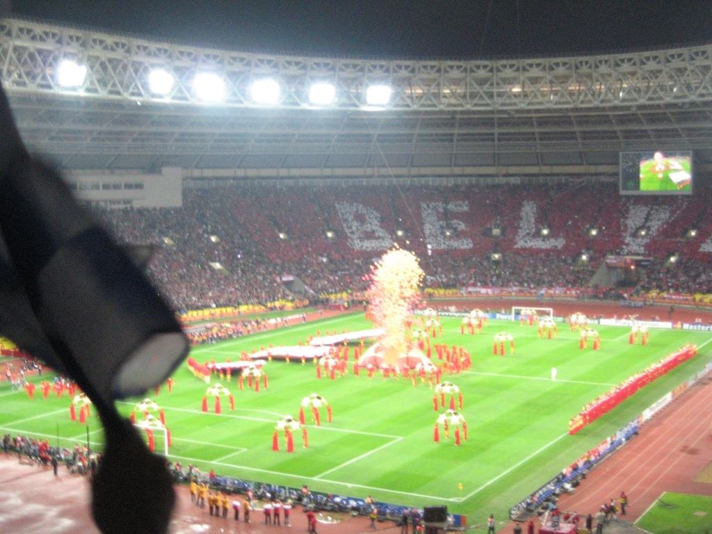 Ein Stadion mit Flutlicht, Eisengittern, Zuschauern, stehenden Personen, Werbetafeln, Anzeigetafeln, Überwachungskamera, Flaggen und Deko-Elementen.