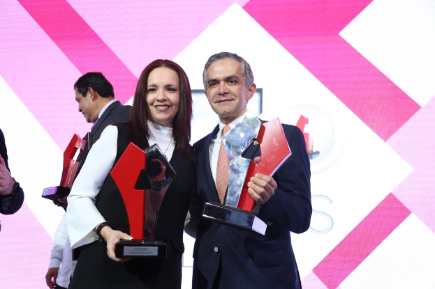 Zwei Personen, die auf einer Bühne lächeln und Trophäen halten, mit einem Bildschirm im Hintergrund.