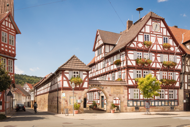 Eine Straße in Rothenburg ob der Tauber, Deutschland, mit alten Gebäuden mit Fenstern, Bäumen, Laternenmasten und einer Person, die ein Fahrrad fährt, mit Hügeln und einer bewölkten Landschaft im Hintergrund.