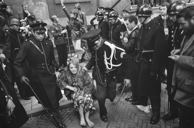 Schwarz-weißes Bild einer Frau, die von der Polizei durch eine Menge von Menschen in Helmen mit Stöcken eskortiert wird, vor einem Gebäude mit Treppe und Geländer im Hintergrund.