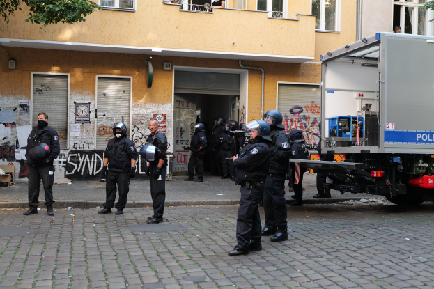 Eine Gruppe von Polizisten in Helmen und mit Waffen steht vor einem Gebäude mit Fenstern, Rollläden und Graffiti, neben einem parkenden Lastwagen und Bäumen im Hintergrund.