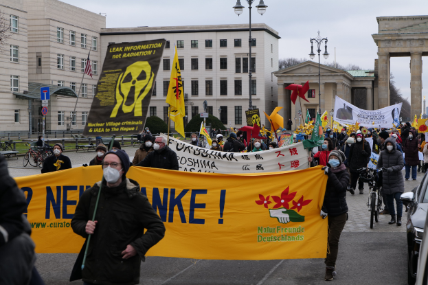 Eine große Gruppe von Menschen marschiert mit Schildern und Fahnen die Straße entlang, mit Fahrzeugen auf der rechten Seite, Gebäuden und Bäumen im Hintergrund und einem Bogen, der unter einem klaren Himmel zu sehen ist, während eine Demonstration gegen Atomkraft in Deutschland stattfindet.