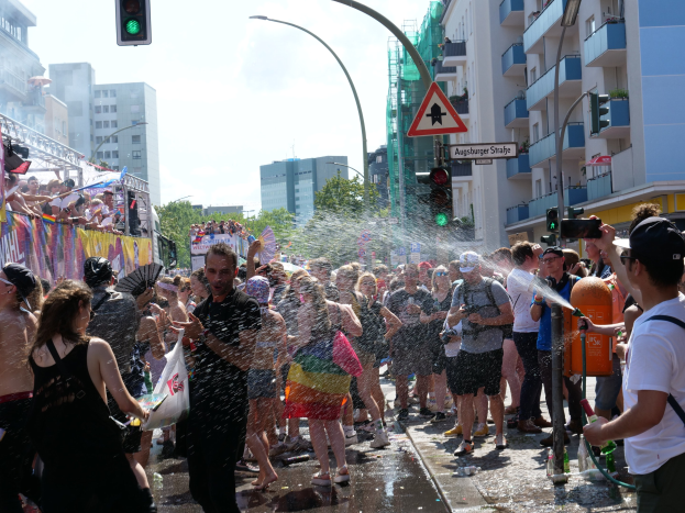 Menschen auf einem Christopher Street Day (CSD) mit Wasser bespritzen sich gegenseitig und halten dabei Gegenstände, mit einem Banner links und Gebäuden, Bäumen und Ampeln im Hintergrund.