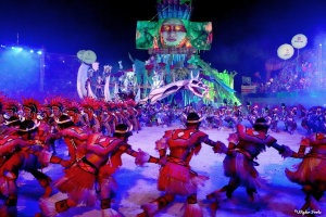 Eine Gruppe von Menschen in farbenfrohen Kostümen tanzt auf einer Bühne vor einem sitzenden Publikum, mit Lichtern, Stangen, Ballons und einem Bildschirm, der eine Person im Hintergrund zeigt, unter einem sichtbaren Himmel, bei der Rio-Karneval 2014.