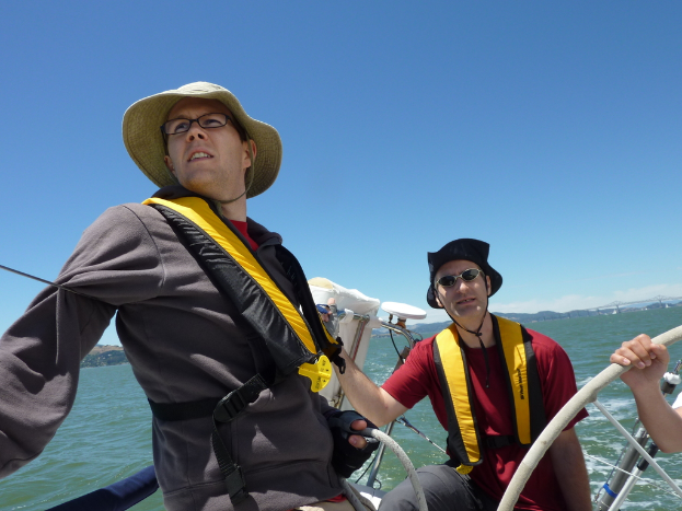 Zwei Männer stehen auf einem Boot, einer trägt einen braunen Hut und ein graues Kleid, der andere einen schwarzen Hut und ein rotes T-Shirt, mit dem Meer im Hintergrund.