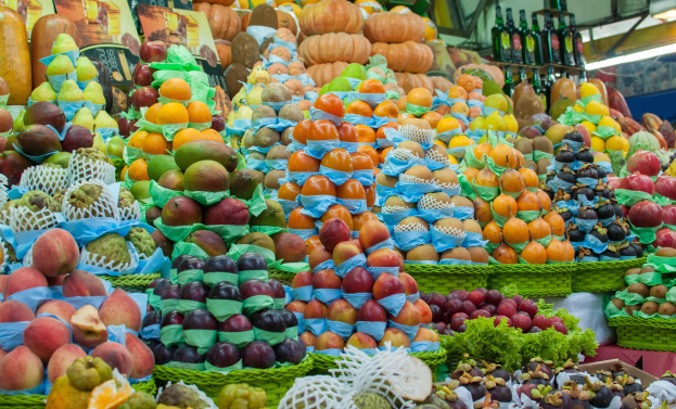 Ein Marktstand mit einer Auswahl an Früchten wie Äpfeln, Orangen und Birnen sowie Gemüse und Flaschen unter Deckenbeleuchtung.