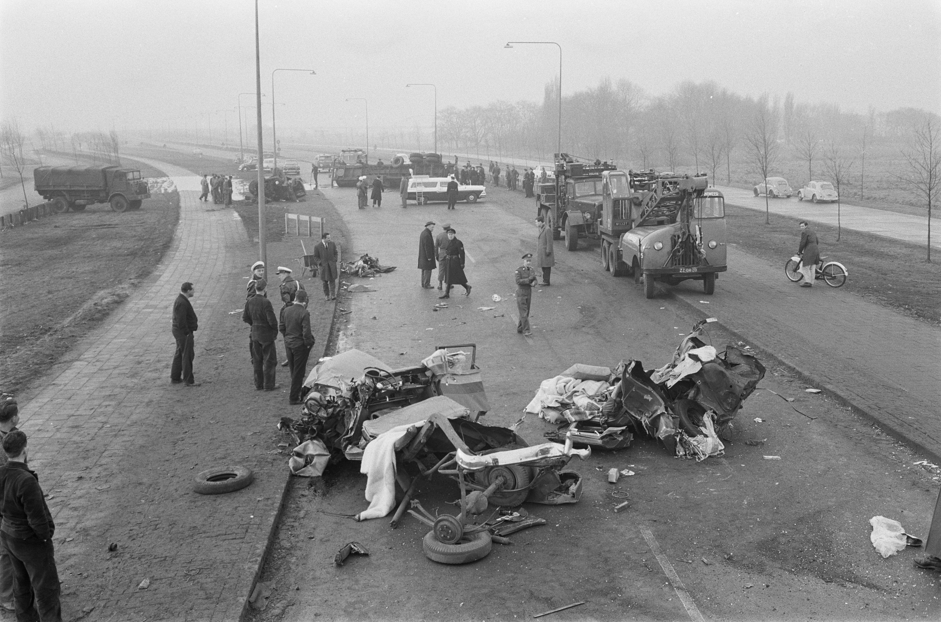 Schwarz-weiß-Szene eines Autounfalls am Straßenrand mit mehreren Fahrzeugen, einer Gruppe von Menschen, Laternenpfählen, Bäumen und dem Himmel im Hintergrund.