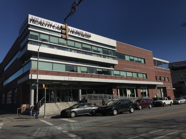 Großes Gebäude mit der Aufschrift "New York City Health Care Homeless Center" mit parkenden Autos, Fußgängern, Straßenmöbeln und Bäumen unter einem klaren Himmel.