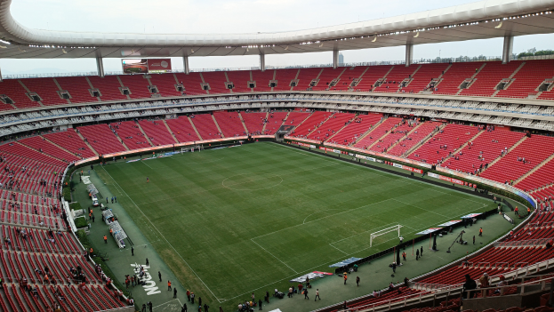 Großes Stadion mit roten Sitzplätzen, Menschen auf dem Feld, ein Tor, Informationsschilder, Flutlicht, ein Bildschirm, Bäume und einen klaren blauen Himmel.