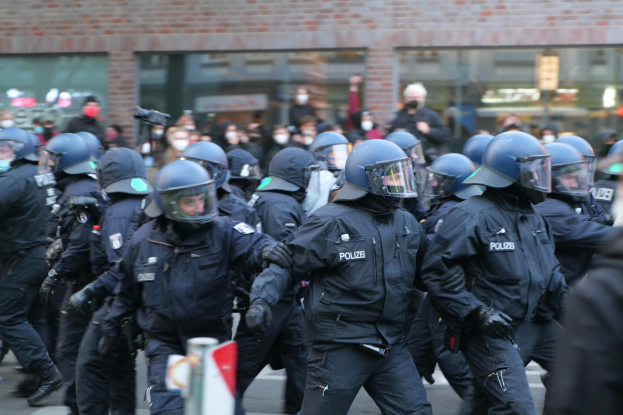 Eine Gruppe von Polizisten in Einsatzausrüstung, einschließlich Helmen und Handschuhen, geht eine Straße entlang mit einem unscharfen Hintergrund, der ein Gebäude mit Glaswänden und ein Schild an einem Pfosten zeigt.