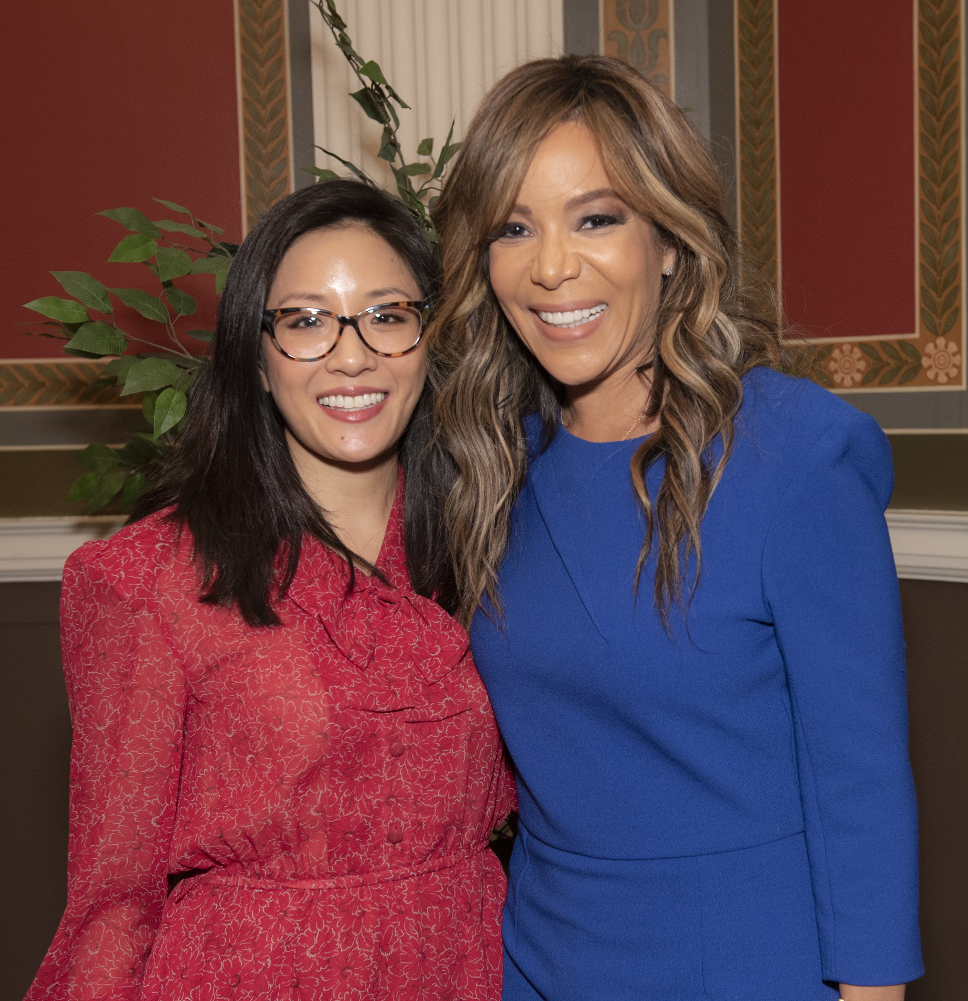 Zwei Frauen in Kleidern stehen lachend nebeneinander auf einem Event, eine in Blau und eine in Rot mit Brille, mit einer Pflanze und einem Vorhang im Hintergrund.
