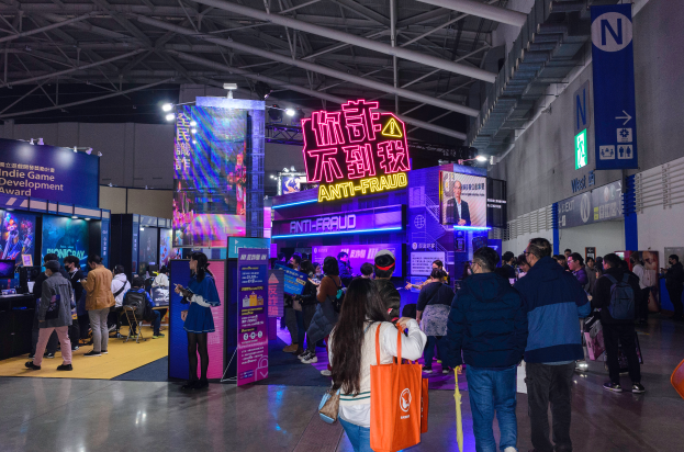 Eine große Gruppe von Menschen steht vor einem Stand auf einer Messe, einige halten Taschen und Schilder mit der Aufschrift "Indie Game Development Award 2019" im Hintergrund.