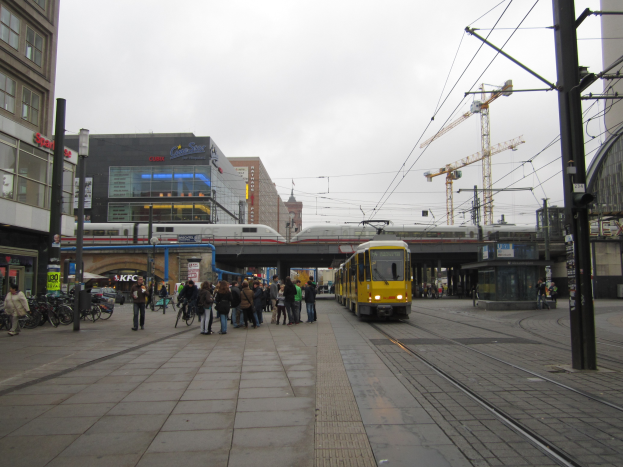 Eine gelbe Straßenbahn auf einer Stadtstraße mit Menschen und Fahrrädern auf dem Gehweg, Masten, Drähte, eine Brücke, einen Zug, Gebäude, Schilder, einen Kran und einen bewölkten Himmel im Hintergrund.
