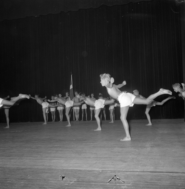 Schwarzes Foto einer Gruppe, die auf einer Bühne mit Vorhang und Fahnenhintergrund Gymnastik macht.