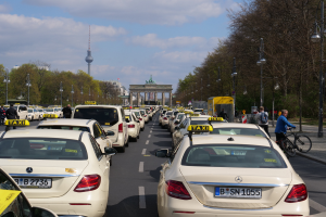 Eine lange Reihe von Taxis parkt am Straßenrand einer belebten Straße in Berlin, Deutschland, mit Fahrzeugen, Radfahrern und Fußgängern, flankiert von Laternen, Bäumen und Gebäuden, einschließlich eines Bogens und eines Turms.
