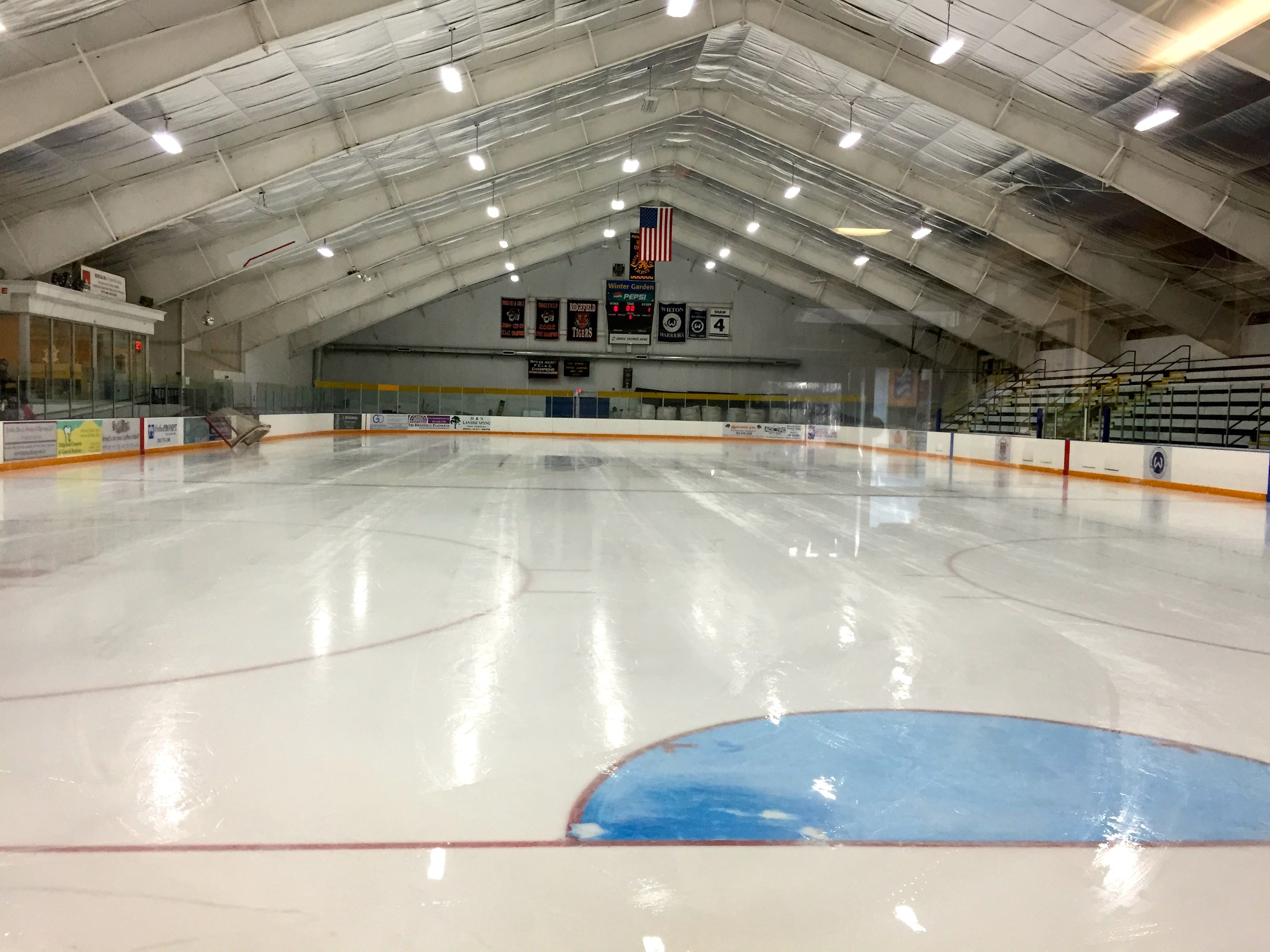 Große Indoor-Eisbahn mit Deckenbeleuchtung, Werbetafeln, einer Rückwand, Treppen mit Geländern und einigen Menschen, die einem Hockeyplatz ähneln.