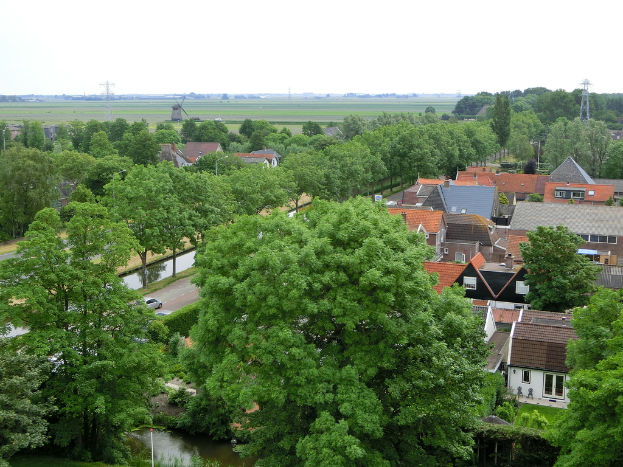 Eine VorstadtSzene mit Häusern und Bäumen, Autos auf einer Straße und Türmen und einer Windmühle im Hintergrund.