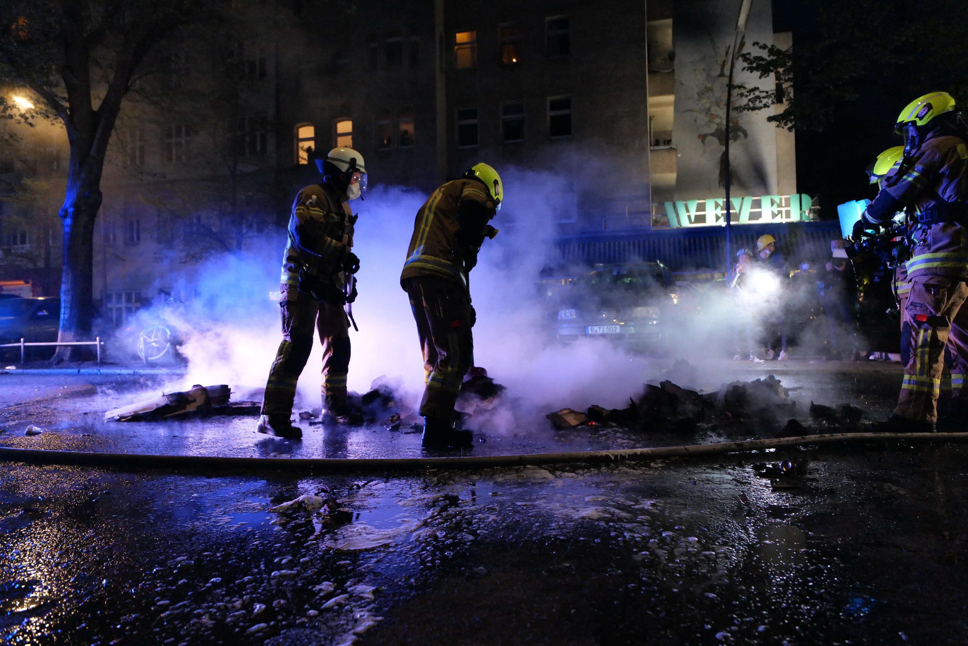 Feuerwehrleute in Helmen stehen nachts auf einer Straße mit Rauch aus einem Rohr, halten Gegenstände, während Fahrzeuge und Gebäude mit beleuchteten Fenstern im Hintergrund erscheinen.
