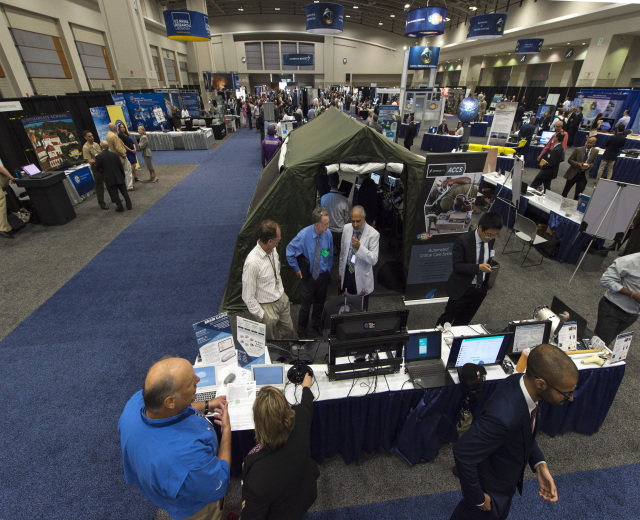 Große Gruppe von Menschen auf einer Karriere-Messe um einen Tisch mit Laptops, Monitoren und Papieren versammelt, mit einem Zelt, Tafeln, Deckenleuchten und Fenstern im Hintergrund.