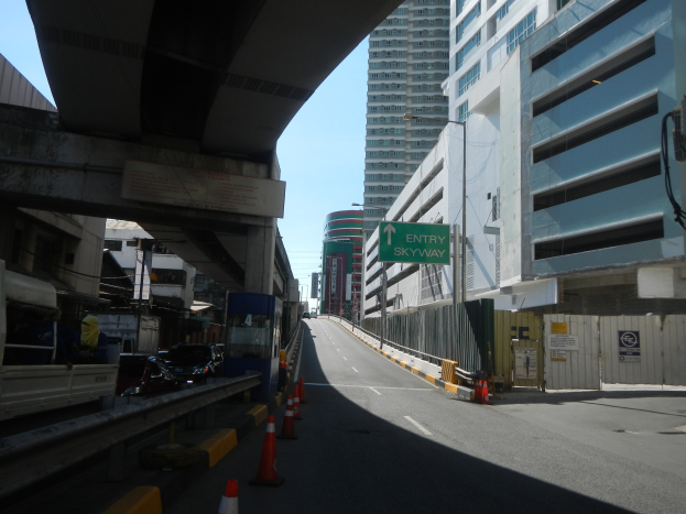 Stadtstraße mit Verkehrskegeln, Fahrzeugen, einer Brücke, Gebäuden mit Namensschildern, Laternen und einem Himmel als Hintergrund.
