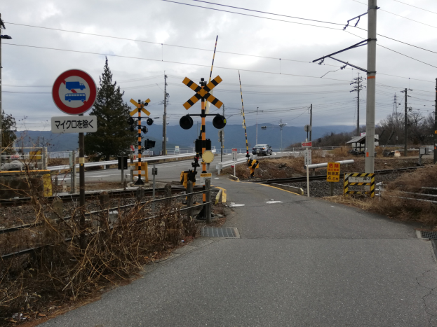 Straße mit einem Bahnübergangsschild, Bahnschienen, Strommasten mit Drähten, Hinweisschildern, Fahrzeugen, Bäumen, Hügeln und einem bewölkten Himmel im Hintergrund.