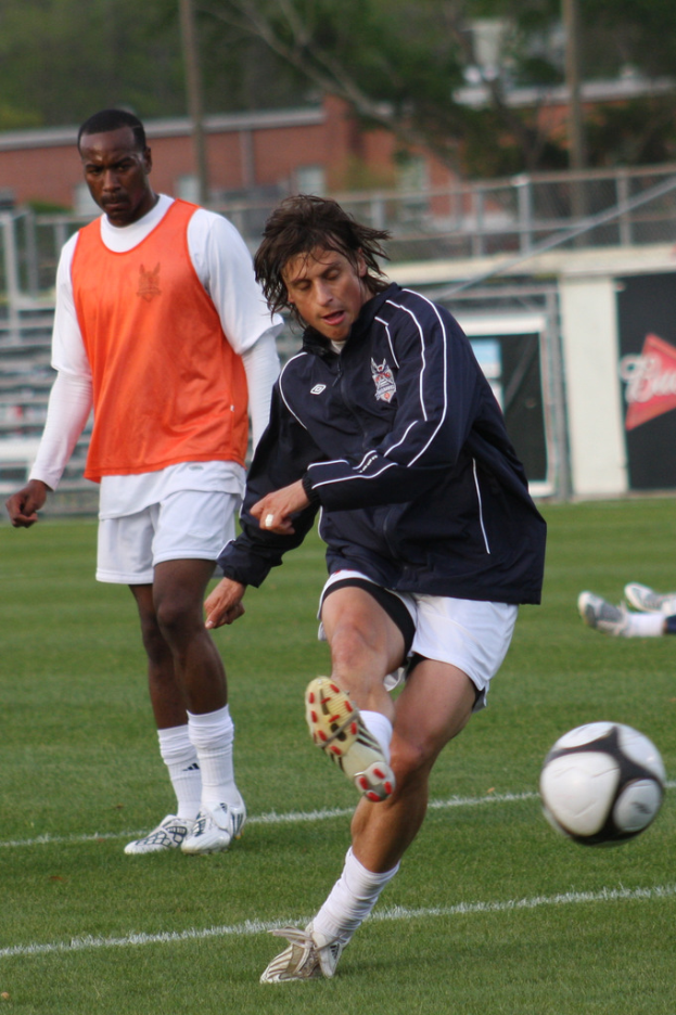 Ein Fussballspieler in einer schwarzen Jacke und weissen Shorts schiesst den Ball, mit einem anderen Spieler in einer orangefarbenen Jacke dahinter und einem Zaun mit Bäumen im Hintergrund.