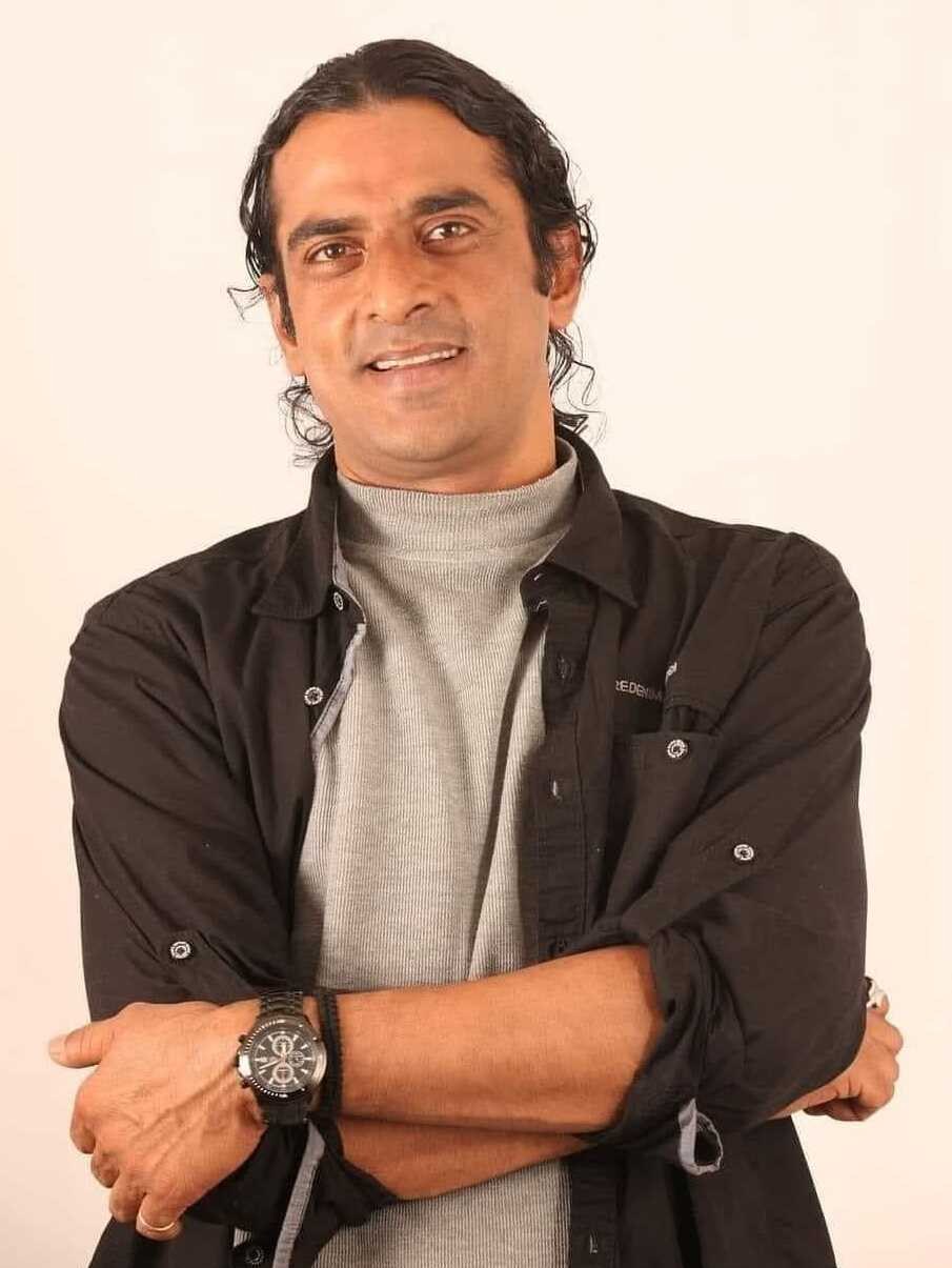 Ein Mann in einem schwarzen und aschfarbenen Kleid mit verschr├Ąckten Armen, der eine Uhr tr├Ągt und gegen einen weißen Hintergrund steht, der in Gedanken versunken scheint.