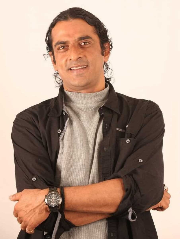 Ein Mann in einem schwarzen und aschfarbenen Kleid mit verschr├Ąckten Armen, der eine Uhr tr├Ągt und gegen einen weißen Hintergrund steht, der in Gedanken versunken scheint.