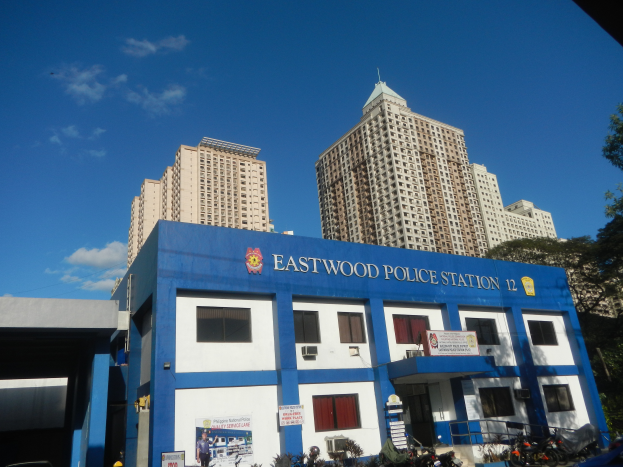 Die Polizeiwache 12 in Manila, Philippinen, ein Gebäude mit Fenstern und Text, umgeben von Pflanzen, geparkten Fahrzeugen, Menschen, einem Schild, Straßenlaternen, Bäumen und unter einem bewölkten Himmel.