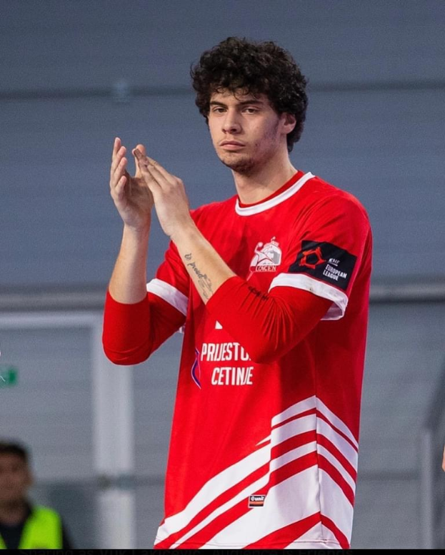 Ein Mann in einem roten und weißen Hemd klatscht in den Hönden in den Vordergrund, mit einer anderen Person und einer Wand im Hintergrund und Text, der unten 'Kopenhagen Futsal-Club - Foto 1' liest.