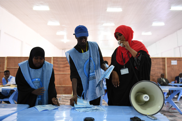 Eine Gruppe von Menschen versammelt sich um einen Tisch mit einem Megafon, Papieren und anderen Gegenständen, die scheinbar eine Wahllokal sind, mit anderen, die im Hintergrund sitzen und stehen, unter beleuchteten Deckeninstallationen.