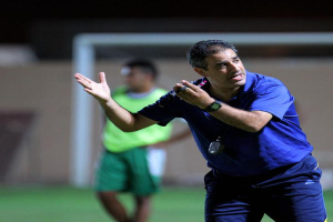 Ein Mann in einem blauen T-Shirt und Hose, der ein Mikrofon hält, steht auf einem Fußballfeld und gestikuliert, mit einer Person in einem grünen Kleid, einem Tor und einer Wand im Hintergrund.