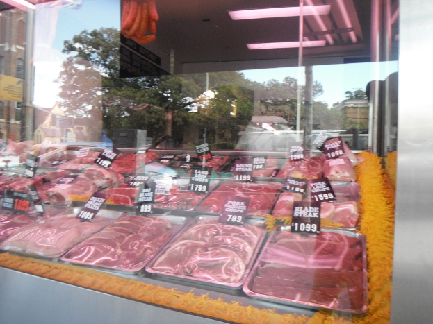 Eine Glasvitrine in einem Fleischgeschäft, die Tabletts mit Fleisch und Preisschildern zeigt, mit Gebäuden und Bäumen im Fenster und der Decke des Raums oben im Bild.
