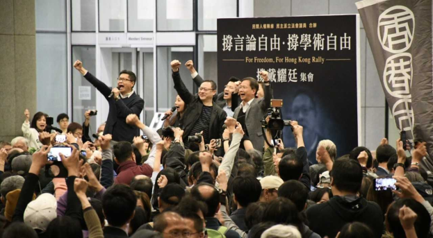 Eine Gruppe von Menschen steht vor einer Menge mit Kameras, Handys und Mikrofonen, mit einem Banner, einer Wand und Glastüren im Hintergrund, was auf eine Protestaktion gegen das Verbot des Hongkonger Auslieferungsgesetzes hindeutet.